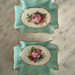 Puede incluir: Dos platos de cerámica turquesa con borde ondulado y ribete dorado. Cada plato presenta un diseño ovalado central con una rosa rosa y un arreglo floral. Los platos se exhiben sobre una superficie de mármol.