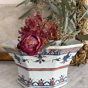 May include: A white, octagonal ceramic vase with blue and orange floral patterns. The vase holds dried flowers and greenery. The vase is sitting on a marble surface.
