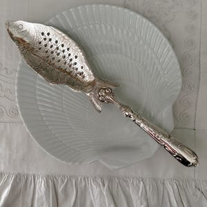 May include: A silver-plated fish-shaped serving utensil with an intricate scale pattern and perforated body. The handle features ornate embossed detailing, connecting to the fish head and tail section, creating a decorative serving tool.