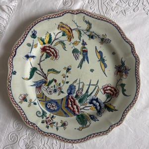 Puede incluir: Un plato crema con borde festoneado con un diseño floral y de pájaros. El diseño pintado a mano presenta flores rojas, amarillas, azules y verdes y un pájaro. El borde tiene un borde rojo y azul.