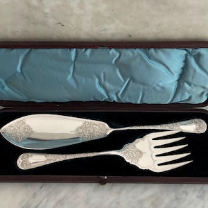 Puede incluir: Un juego de servicio de plata con un cuchillo y un tenedor en una caja forrada de terciopelo marrón. El juego tiene un diseño ornamentado.