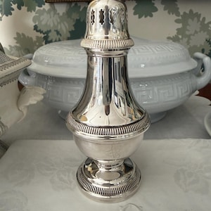 Puede incluir: Una lámpara de aceite de plata pulida con una parte superior decorativa calada. La lámpara tiene un cuerpo bulboso y una base acanalada. La superficie reflectante de la lámpara muestra los objetos circundantes. Es un artículo vintage.