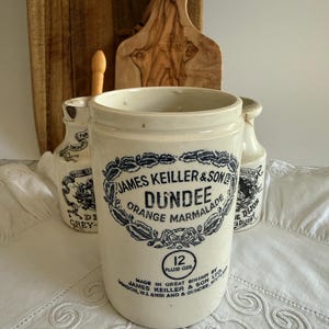 May include: A cylindrical off-white ceramic jar featuring black printed text that reads James Keiller & Son Ltd Dundee Orange Marmalade, with a 12 fluid ozs label, surrounded by two smaller ceramic jars and a wooden board in the background.
