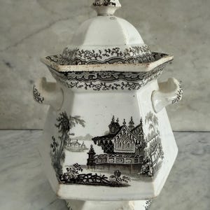 Puede incluir: Jarra con tapa de cerámica blanca y negra con un diseño detallado de paisaje. La jarra tiene forma hexagonal con asas y un adorno decorativo en la tapa. El paisaje presenta edificios, árboles y un puente.