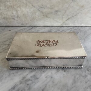 May include: A rectangular silver box with a decorative rope-like border. The lid features an engraved monogram in a cursive font. The box is displayed on a marble surface.