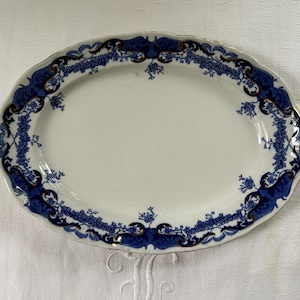 May include: An oval ceramic serving platter with a cream-coloured centre and a decorative border featuring deep blue floral patterns, intricate scrollwork, and subtle gold-toned accents along the scalloped edge of the rim.
