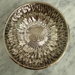 Puede incluir: Un cuenco redondo de plata con una superficie interior martillada y texturizada. El centro del cuenco presenta un monograma ornamentado grabado que consiste en las letras entrelazadas LR. El acabado metálico tiene un aspecto pulido y reflectante.