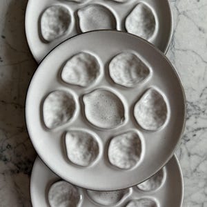 May include: Three white ceramic oyster plates with a textured surface and a dark rim. Each plate has eight individual oyster shell-shaped indentations. The plates are stacked on top of each other.