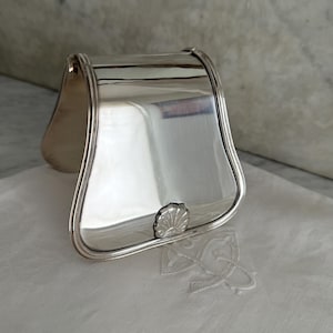 May include: A polished silver napkin holder with a shell-shaped accent. The holder has a curved top and a rectangular base, reflecting the surrounding environment. The napkin holder is resting on a white surface.