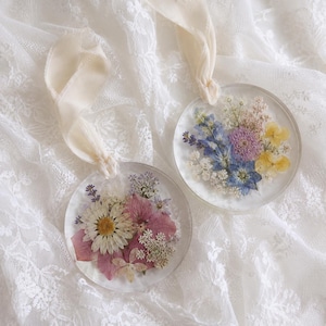 May include: Two circular clear resin ornaments featuring embedded pressed flowers. One ornament contains pink and white daisies with lavender, while the other displays blue, yellow, and purple blossoms. Each piece hangs from a cream-colored silk ribbon.