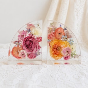 May include: Two semi-circular clear resin bookends containing preserved flowers. The left piece features deep pink, light pink, and white roses. The right piece displays orange, yellow, and light pink roses with small blue and purple accent flowers.