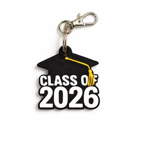 May include: A black graduation cap keychain with the text "CLASS OF 2026" in white. The tassel is yellow. The keychain has a silver clasp and ring.