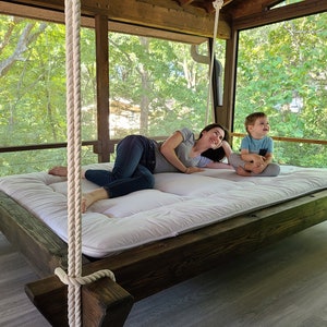 May include: A white hanging bed with a wooden frame and rope supports. A woman and a child are relaxing on the bed, which is suspended from the ceiling of a screened porch.