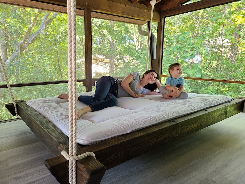May include: A white hanging bed with a wooden frame and rope supports. A woman and a child are relaxing on the bed, which is suspended from the ceiling of a screened porch.