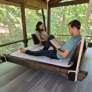 May include: A family of three relaxes on a wooden swing bed, reading books on a screened-in porch. The swing bed is suspended from the ceiling by thick ropes. The porch has a view of a lush green forest.