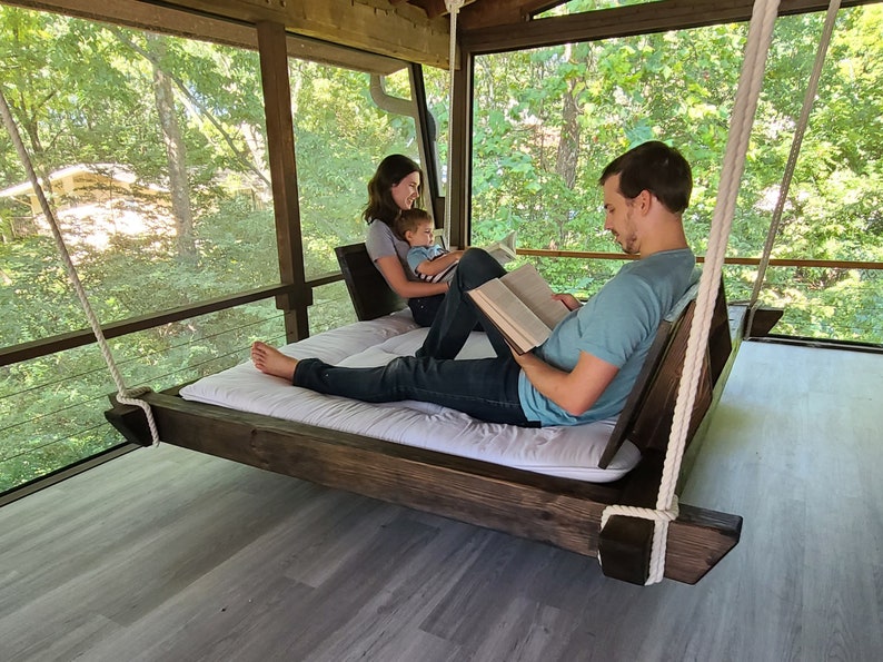 May include: A family of three relaxes on a wooden swing bed, reading books on a screened-in porch. The swing bed is suspended from the ceiling by thick ropes. The porch has a view of a lush green forest.