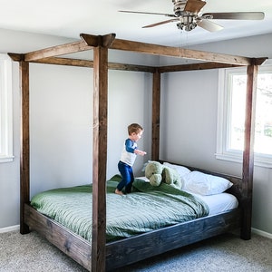 May include: A wooden canopy bed frame with a green duvet and white pillows. A young child is jumping on the bed.