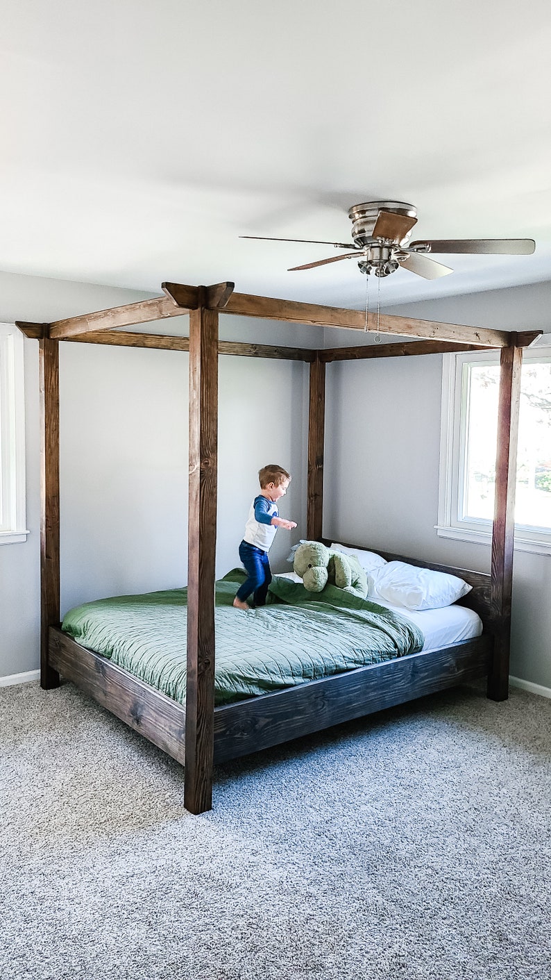 May include: A wooden canopy bed frame with a green duvet and white pillows. A young child is jumping on the bed.