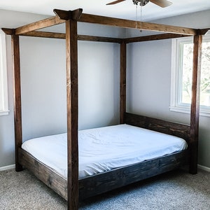 May include: A wooden canopy bed frame with a white mattress. The bed frame is made of dark brown wood and has a simple design. The bed is in a room with a ceiling fan and carpeted floors.