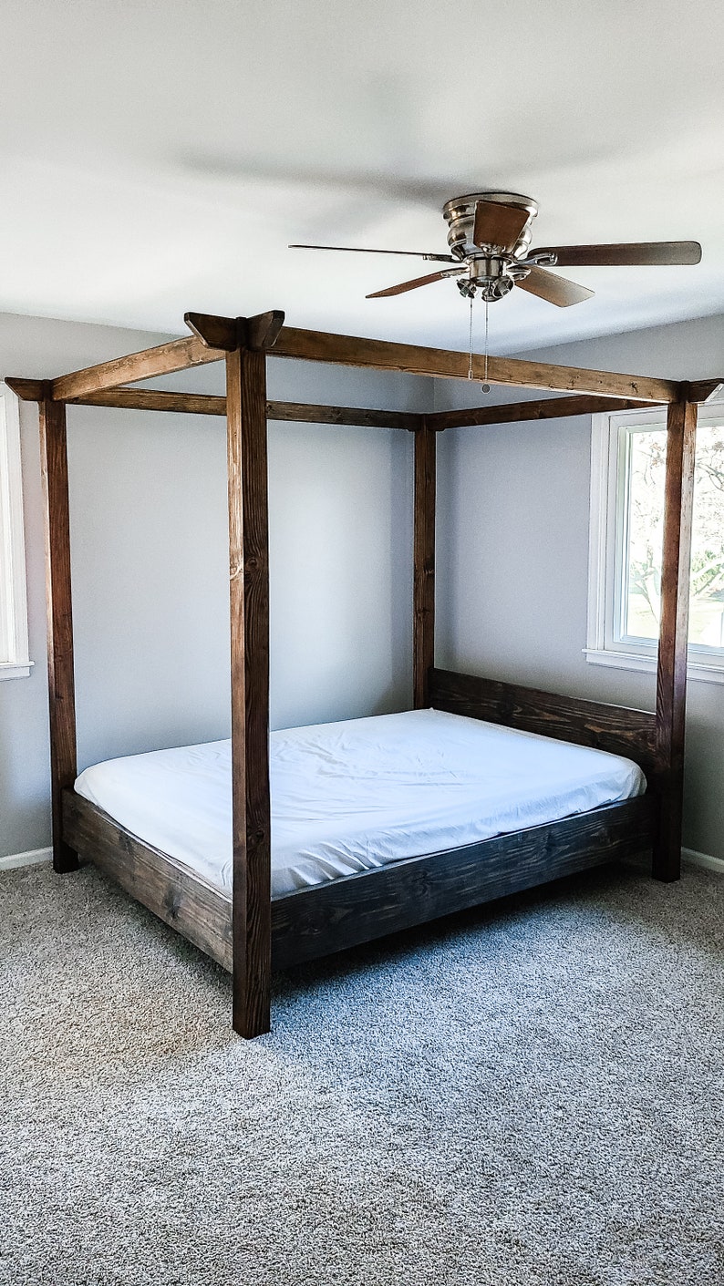 May include: A wooden canopy bed frame with a white mattress. The bed frame is made of dark brown wood and has a simple design. The bed is in a room with a ceiling fan and carpeted floors.