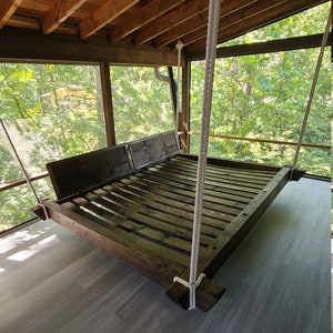 May include: A wooden swing bed hangs from the ceiling of a screened-in porch. The bed is made of dark wood and has a slatted design. The porch has a wooden floor and a view of trees outside.