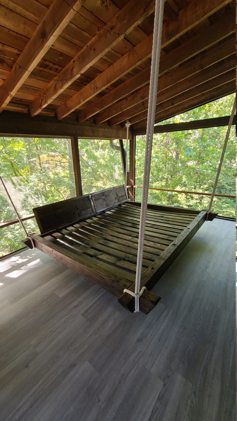 May include: A wooden swing bed hangs from the ceiling of a screened-in porch. The bed is made of dark wood and has a slatted design. The porch has a wooden floor and a view of trees outside.