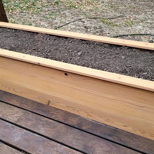 May include: A long, rectangular wooden planter box filled with dark brown soil. The planter is made of natural wood with a light brown finish. It is sitting on a wooden deck.