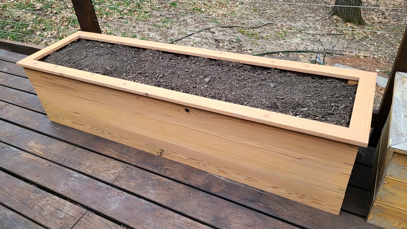 May include: A long, rectangular wooden planter box filled with dark brown soil. The planter is made of natural wood with a light brown finish. It is sitting on a wooden deck.