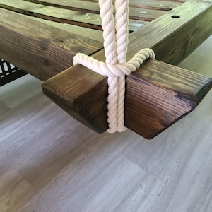 May include: Close-up of a wooden swing frame with a white rope attached. The rope is tied in a knot and the frame is stained a dark brown color.