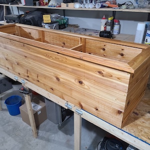 May include: A long, rectangular wooden planter box with a natural wood finish. The planter is made of cedar wood and has a raised edge around the top. The planter is sitting on a wooden workbench.