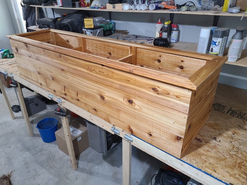 May include: A long, rectangular wooden planter box with a natural wood finish. The planter is made of cedar wood and has a raised edge around the top. The planter is sitting on a wooden workbench.