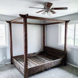 May include: A dark brown wooden canopy bed frame with a slatted headboard and footboard. The bed frame is in a bedroom with a ceiling fan and carpeted floors.