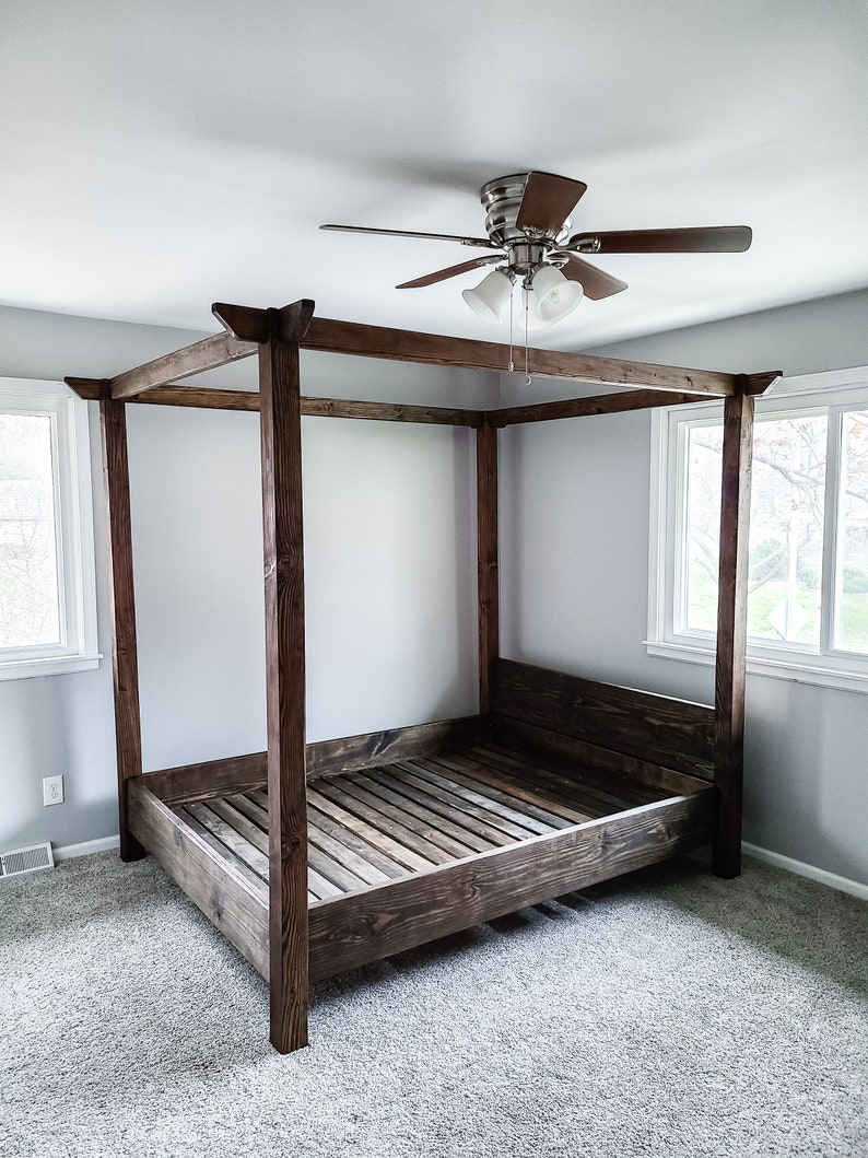 May include: A dark brown wooden canopy bed frame with a slatted headboard and footboard. The bed frame is in a bedroom with a ceiling fan and carpeted floors.