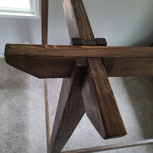 May include: Close-up of a wooden bed frame with a dark brown stain. The frame is made of thick, sturdy wood and features a unique design with angled legs and a cross-beam.