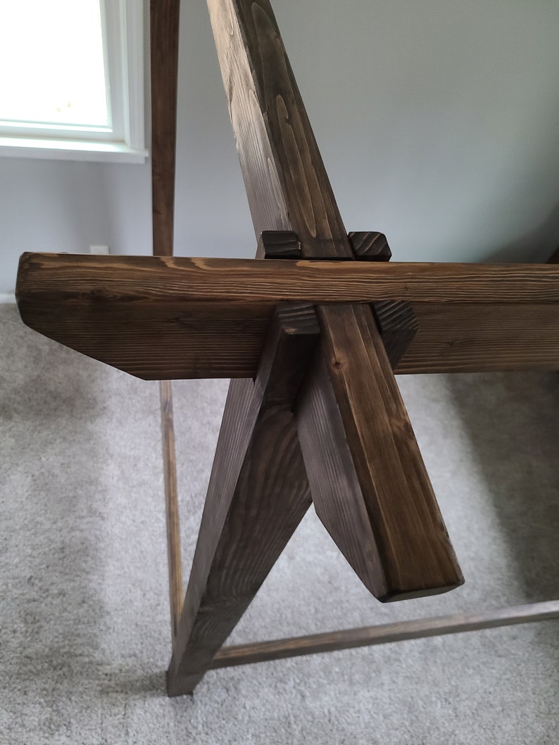 May include: Close-up of a wooden bed frame with a dark brown stain. The frame is made of thick, sturdy wood and features a unique design with angled legs and a cross-beam.