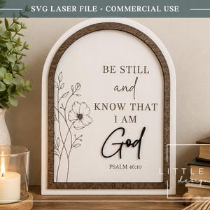 Może przedstawiać: Łukowaty drewniany znak z białym tłem zawierający czarny tekst BE STILL and KNOW THAT I AM God oraz PSALM 46:10. Projekt zawiera czarną ilustrację kwiatową wykonaną liniami po lewej stronie oraz ciemnobrązową drewnianą ramę.