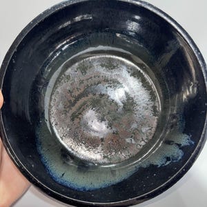 May include: A handmade ceramic bowl with a dark, glossy exterior. The interior features a metallic, speckled finish with hints of blue and green. The bowl's rim is dark, and the overall design is simple and elegant.