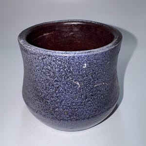 May include: A handmade ceramic cup with a speckled blue exterior and a deep burgundy interior. The cup has a rounded base and a slightly flared rim. The texture of the cup appears rough and organic, suggesting a handcrafted process.