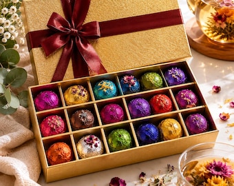 May include: A rectangular gold gift box with a dark red satin ribbon bow containing fifteen spherical tea balls wrapped in colorful metallic foil. Each tea ball is topped with a small dried flower petal, arranged in a grid inside the box.