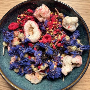 May include: A teal ceramic bowl filled with a colorful mix of dried ingredients. The mix includes vibrant blue cornflowers, red berries, and white, textured pieces. The bowl sits on a light brown wooden surface.