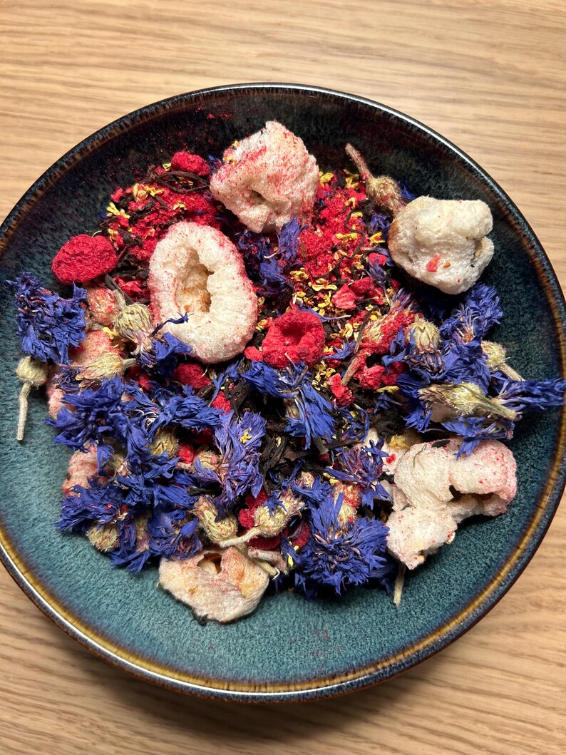 May include: A teal ceramic bowl filled with a colorful mix of dried ingredients. The mix includes vibrant blue cornflowers, red berries, and white, textured pieces. The bowl sits on a light brown wooden surface.