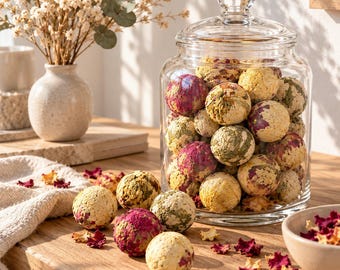 May include: A clear glass apothecary jar with a lid filled with spherical bath bombs decorated with dried flower petals in shades of pink, yellow, and green. Several loose bath bombs and scattered dried flower petals rest on the wooden surface.