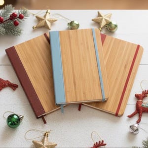 May include: Three bamboo notebooks with colored accents, including red and blue, are arranged on a white surface. Christmas ornaments, such as gold stars, green baubles, and red reindeer, surround the notebooks, creating a festive holiday theme.