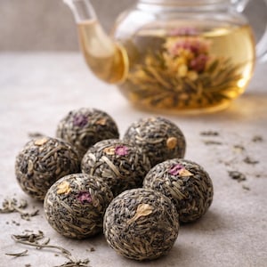 Puede incluir: Una tetera de vidrio transparente llena de té dorado, mostrando una flor floreciente. Siete bolas de té, envueltas en hojas de té verde con pétalos de flores visibles, están dispuestas al frente. Hojas de té sueltas esparcidas.