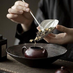 May include: A person pouring loose leaf tea from a silver scoop into a small, maroon teapot. The teapot sits in a dark, shallow bowl. The scene is set on a brown surface, with a black background.