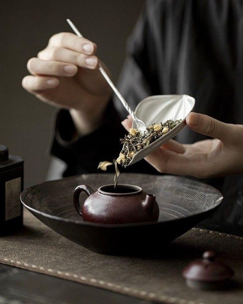 May include: A person pouring loose leaf tea from a silver scoop into a small, maroon teapot. The teapot sits in a dark, shallow bowl. The scene is set on a brown surface, with a black background.