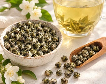 May include: A ceramic bowl filled with tightly rolled green tea pearls, accompanied by a wooden scoop containing more pearls, a glass cup of brewed tea with visible leaves, and fresh white jasmine flowers with green leaves.
