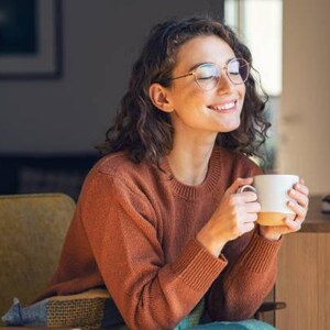 May include: A person wearing glasses and a rust-colored sweater smiles while holding a white mug with a tan base. The person is seated in a yellow armchair, with a dark background and a wooden table visible.
