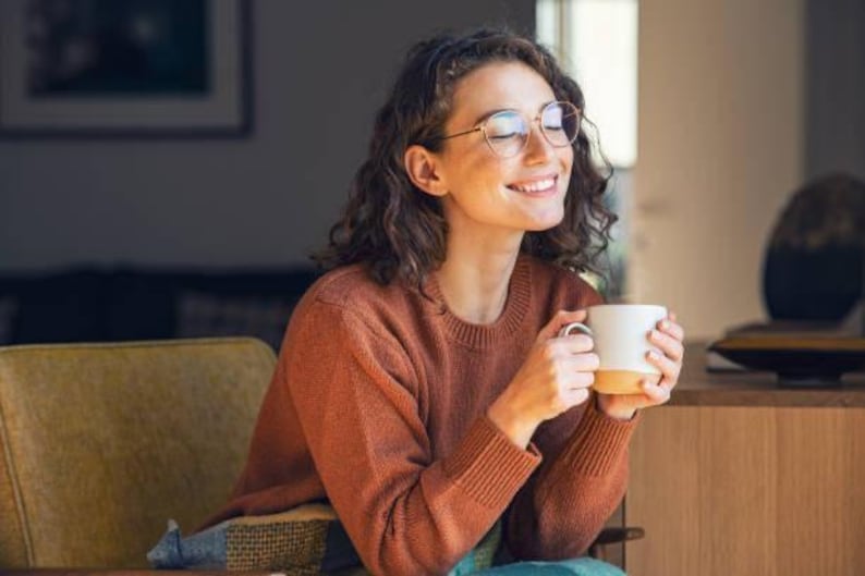 May include: A person wearing glasses and a rust-colored sweater smiles while holding a white mug with a tan base. The person is seated in a yellow armchair, with a dark background and a wooden table visible.