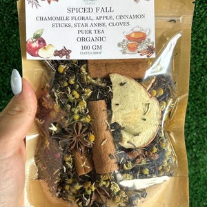 May include: A clear bag filled with organic Spiced Fall Puer tea. The tea blend includes chamomile, apple, cinnamon sticks, star anise, and cloves. The bag has a label with the text "Spiced Fall" and "100 GM".